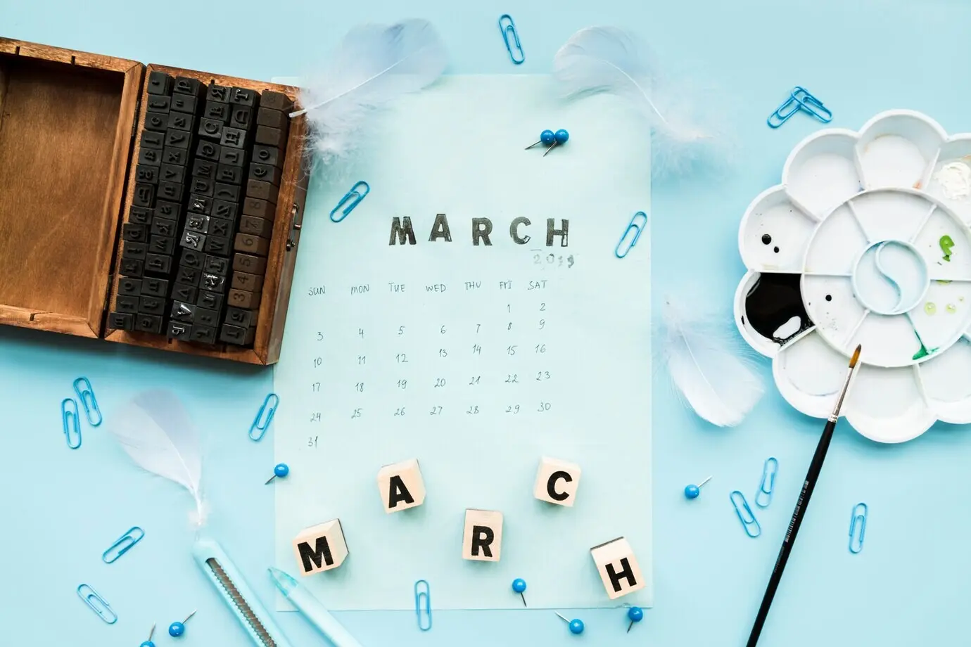 Wooden typographic blocks, a feather, March blocks and a March stamp on a calendar with stationery, set against a blue backdrop.